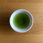 Saemidori 2026 sencha brewed in a small white Japanese cup on a wooden table, top-down view — vivid bright green liquor