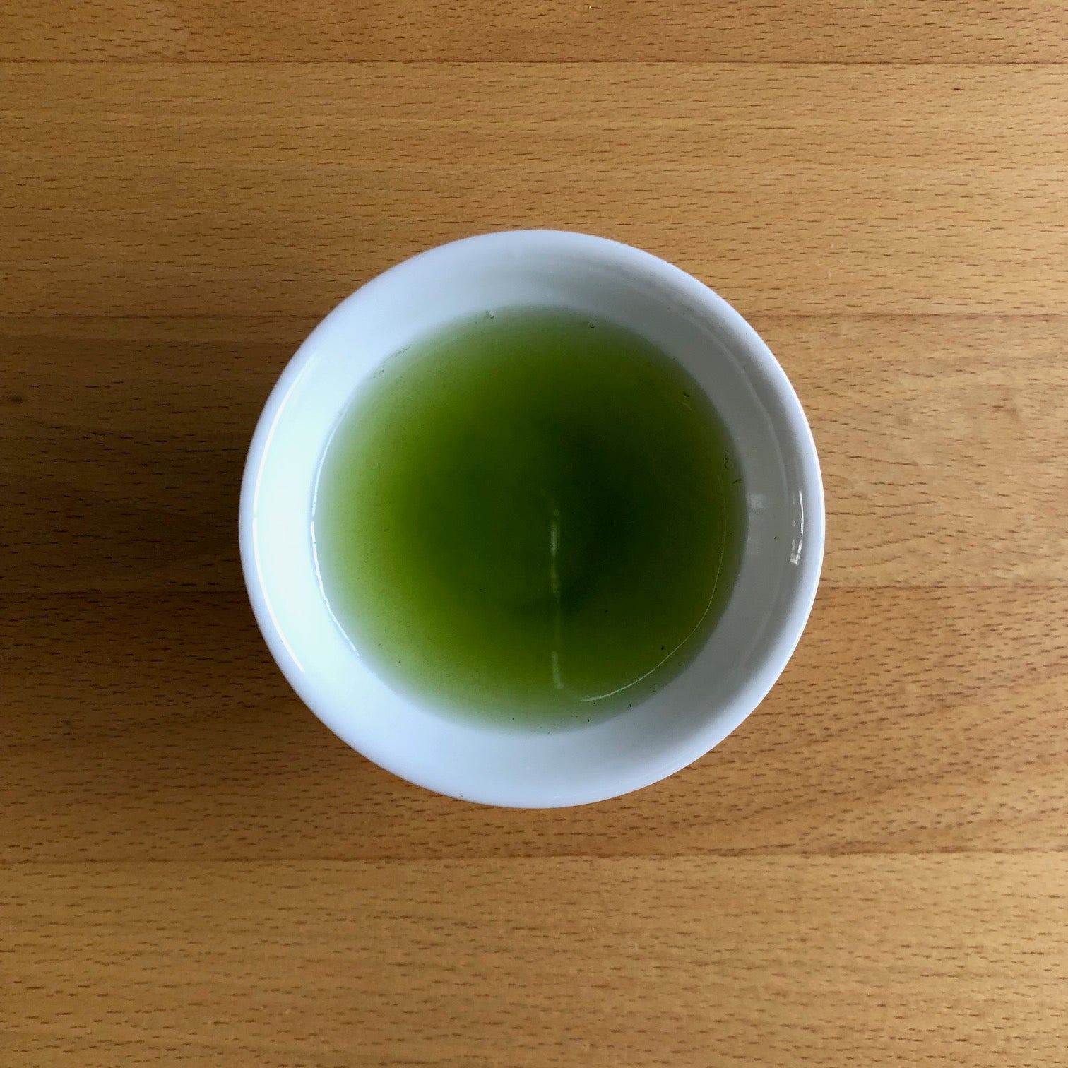 Saemidori 2026 sencha brewed in a small white Japanese cup on a wooden table, top-down view — vivid bright green liquor