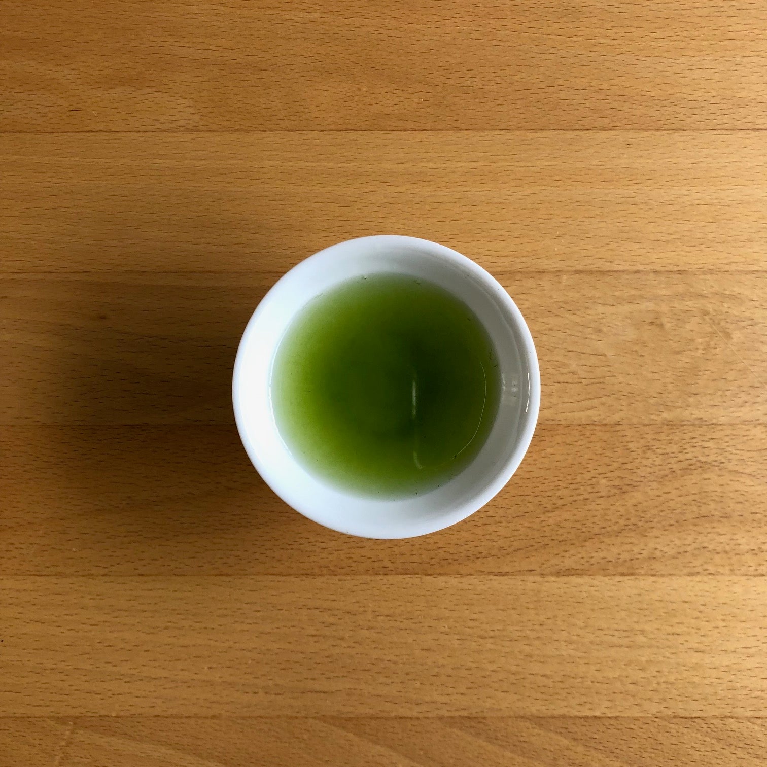 Saemidori 2026 sencha brewed in a small white Japanese cup, photographed from above on a wooden table — vivid bright green liquor