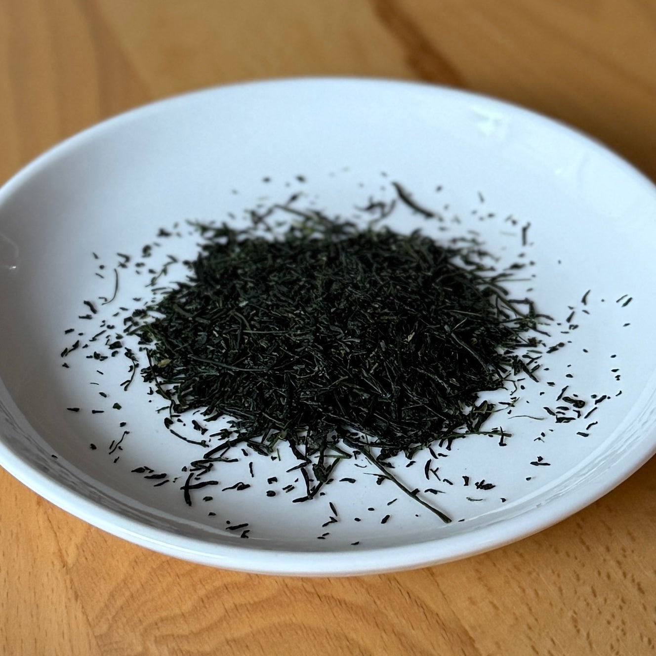 Dried Hoshino Shizuku 2026 sencha leaves on a white plate, top-down view — fine dark-green needle-shaped leaves