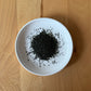 Dried Hoshino Shizuku 2026 sencha leaves on a white plate, top-down view — smaller heap, lighter spread